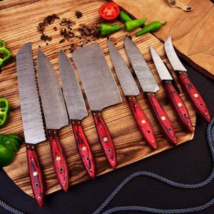 May include: A set of eight kitchen knives with stainless steel blades and red wooden handles. The knives are arranged on a wooden cutting board, with a tomato and green peppers in the background. The blades have a unique pattern.