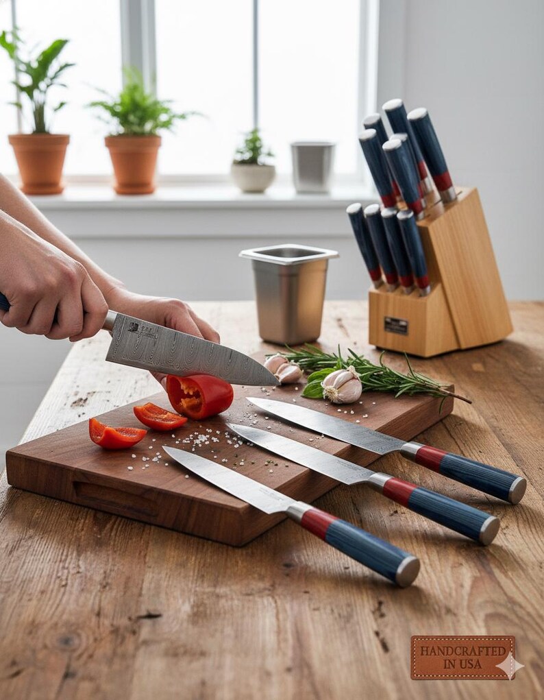 Puede incluir: Una tabla de cortar de madera con un pimiento rojo cortado por un cuchillo de acero inoxidable. Tres cuchillos adicionales con mangos azules y rojos est&aacute;n en la tabla. Un bloque de cuchillos de madera contiene m&aacute;s cuchillos. La imagen est&aacute; etiquetada "Handcrafted in USA."
