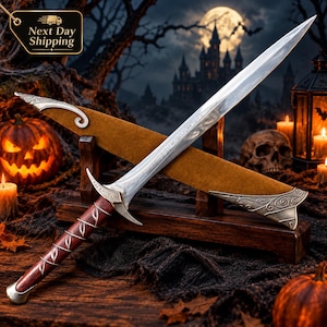May include: A silver-toned metal sword with a pointed blade featuring intricate engravings, a dark wood handle with silver leaf-like inlays, and a matching brown scabbard with ornate silver metal accents, all resting on a wooden display stand.