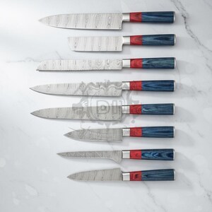 Puede incluir: Un juego de siete cuchillos de cocina de acero inoxidable con hojas estampadas y mangos rojos, plateados y azules. Los cuchillos est&aacute;n colocados sobre una superficie de m&aacute;rmol blanco. El texto "HANDCRAFTED IN USA" est&aacute; en la parte inferior.