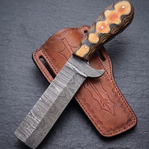 May include: A Damascus steel knife with a patterned blade and a wooden handle with orange accents. The knife rests in a brown leather sheath with decorative stitching and an arrow design.
