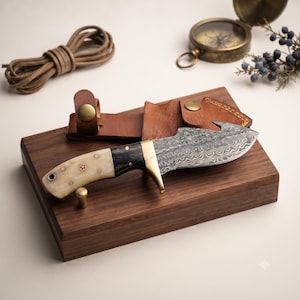 May include: A fixed-blade knife with a Damascus steel blade featuring a gut hook, a brass guard, and a two-tone handle made of light-colored bone and dark wood. The knife is accompanied by a brown leather sheath with brass snap closures.