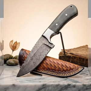 Puede incluir: Un cuchillo de hoja fija con una hoja de acero de Damasco con diseño ondulado, un mango gris oscuro con tres remaches plateados y una funda de cuero marrón con textura trenzada y costuras amarillas visibles en los bordes.