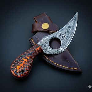 Puede incluir: Un cuchillo karambit de acero de Damasco con un mango con diseño naranja y negro, en una funda de cuero marrón. La hoja curva tiene un agujero circular cerca del mango. La funda tiene un lazo para cinturón y un botón dorado.