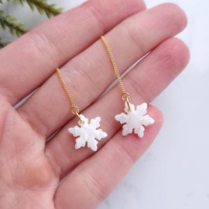 May include: A pair of delicate snowflake earrings. Each earring features a white, snowflake-shaped charm suspended from a fine gold chain. The earrings are held in a hand, with a blurred green plant in the background.