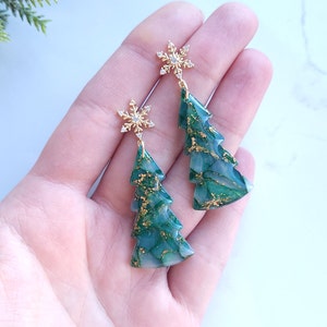 May include: Dangle earrings featuring green Christmas tree shapes with gold accents. Each earring is topped with a gold snowflake embellished with small clear crystals. The earrings are a festive holiday accessory.