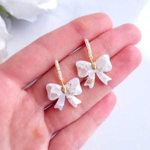 Puede incluir: Un par de pendientes blancos con forma de lazo con detalles dorados y aros con perlas. Los pendientes se sostienen en una mano, mostrando su delicado diseño y detalles intrincados. Los lazos tienen una apariencia texturizada con motas doradas.