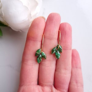 May include: A pair of gold earrings with green leaf-shaped charms. The leaves have a marbled pattern and are attached to a gold hoop.