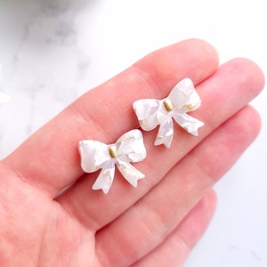 Könnte beinhalten: Ein Paar weiße Schleifenohrringe mit goldenen Akzenten. Die Schleifen bestehen aus einem weißen Material mit goldenen Flocken.