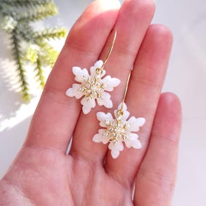 May include: A pair of snowflake earrings. Each earring has a white, six-pointed snowflake design with a cluster of small, sparkling crystals at the centre. The earrings hang from delicate gold-coloured hoops.