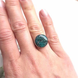 May include: A silver ring with a round, turquoise-coloured stone set in the centre. The stone has a speckled pattern.