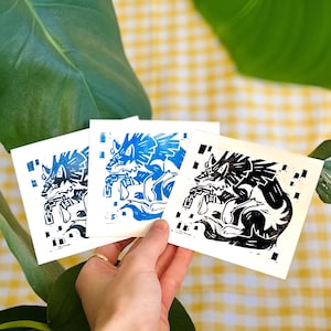 Peut inclure: Trois estampes en linogravure noir et blanc d'une créature ressemblant à un loup avec des ailes, portant une chaîne autour du cou. Les estampes sont sur du papier blanc et sont légèrement différentes en taille.