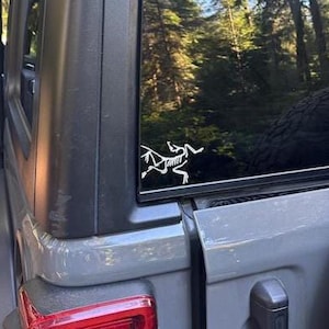 May include: A white vinyl decal of an Archaeopteryx fossil skeleton applied to the rear side window of a grey SUV. The decal features the distinct skeletal structure of the prehistoric bird, positioned against the dark tinted glass of the vehicle.