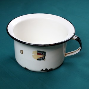 May include: White enamel chamber pot with a black rim and handle. The pot has a small, faded emblem on the side. The interior is white, and the handle is black and white. The pot is on a teal surface.