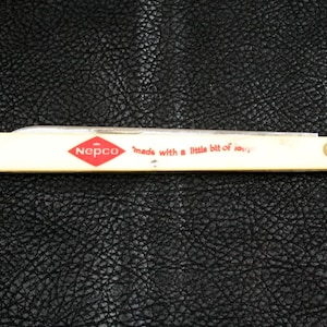 May include: A vintage folding knife with a cream-colored handle. The handle features the red diamond-shaped "Nepco" logo and the text "made with a little bit of love" in red. The knife is closed and resting on a textured black surface.