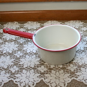 May include: White enamel saucepan with a red rim and a red handle. The saucepan is sitting on a white crocheted doily.