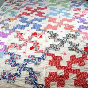 May include: A handmade quilt with a repeating pattern of colorful, interlocking shapes in red, blue, purple, and floral prints. The quilt is on a bed and has pink pom-poms attached at intervals. The overall color is white.
