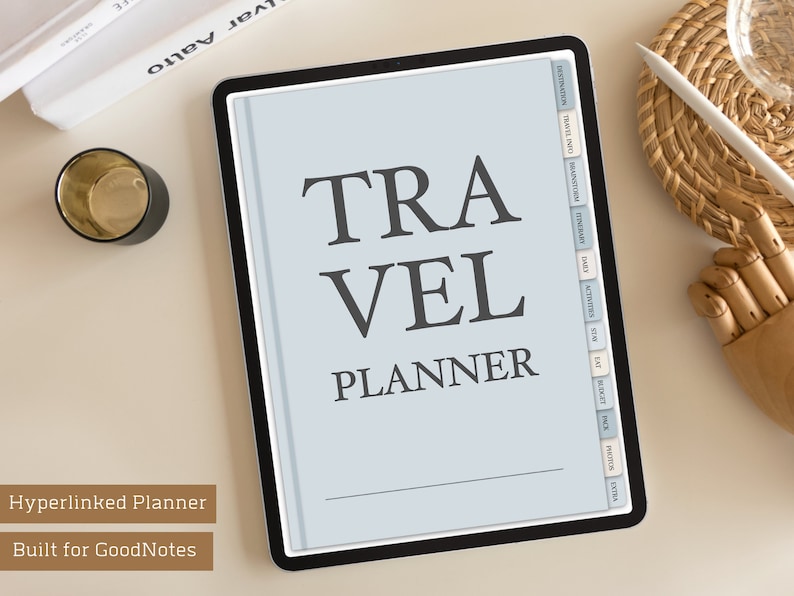 May include: A digital tablet displaying a travel planner template with the text "TRAVEL PLANNER" in black font on a light blue background. The tablet has a black frame and is on a white surface. The template has tabs for different sections of the planner, including "Destination", "Travel Info", "Brainstorm", "Itinerary", "Dates", "Activities", "Stop", "Est", "Budget", "Pack", "Photos", and "Extra".