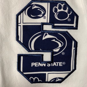 Pode incluir: Um remendo bordado em azul marinho e branco com as palavras "Penn State" e um logotipo de leão estilizado. O remendo é rodeado por uma borda azul marinho.