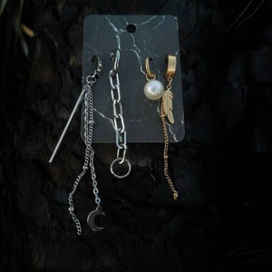 May include: Three silver and gold ear cuffs with dangling chains. The chains have a crescent moon, a pearl, and a feather charm.