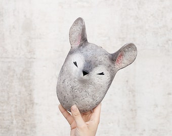 Può includere: Una maschera di animale in ceramica grigia con gli occhi chiusi e un piccolo naso nero. La maschera è tenuta in una mano con un tatuaggio nero.
