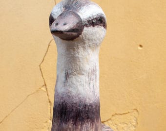 May include: A handcrafted, decorative bird sculpture with a long neck and rounded body. The bird features a color palette of white, brown, and purple, with a textured, wood-like appearance. The sculpture is held in front of a textured, yellow wall.