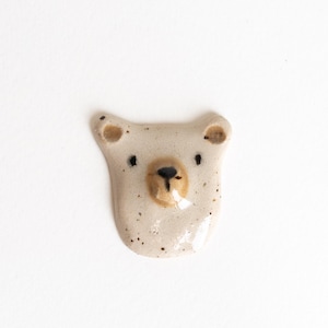 Könnte beinhalten: Ein kleines, cremefarbenes Keramik-Bärenkopf-Ornament mit glänzender Oberfläche und dunkelbraunen Sprenkeln. Der Bär hat eine hellbraune Schnauze, eine schwarz bemalte Nase und einen Mund sowie zwei kleine schwarze Augen.