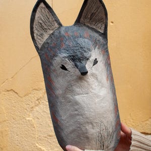 May include: A handmade, paper mache fox mask with a whimsical design. The mask features a gray and white color scheme with red accents, black ears, and a black nose. The fox mask is held in front of a yellow wall.