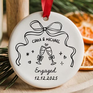 May include: A white ceramic ornament with a red ribbon, featuring a black line drawing of champagne glasses, a bow, and the text "Ciara & Michael Engaged 12.12.2025." The ornament is on a wooden tray with a lit candle and dried orange slices.