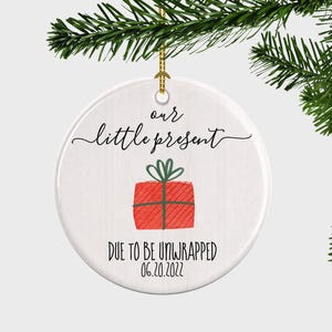 May include: White ceramic Christmas ornament with the text "our little present" and "DUE TO BE UNWRAPPED 06.20.2022". The ornament features a red gift with a green bow. A gold string is attached for hanging.