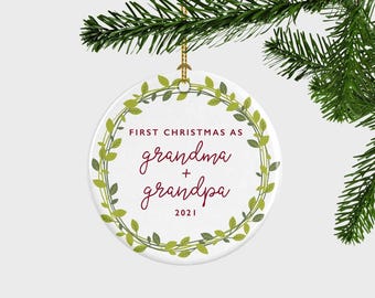 Peut inclure: Une décoration ronde en céramique blanche ornée d'une couronne de feuilles vertes. Le texte FIRST CHRISTMAS AS grandma + grandpa 2021 est inscrit en rouge foncé. L'ornement est suspendu par un cordon doré à une branche de pin.
