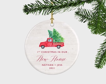May include: A round white ceramic ornament with a wood grain texture featuring a red truck carrying a green pine tree. The text reads 1ST CHRISTMAS IN OUR New Home NATHAN + JESS 2021. It hangs from a gold string against a pine branch.