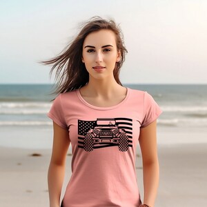 May include: A pink t-shirt featuring a black graphic of a jeep driving over an American flag.