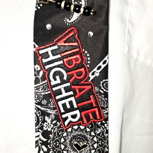 Peut inclure: Cravate noire à motif cachemire avec l'inscription brodée "VIBRATE HIGHER" en rouge et blanc. La cravate est ornée de petits strass transparents et d'une épingle décorative avec des perles noires et des détails dorés. La cravate est présentée sur une chemise blanche.