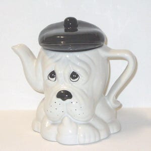 Könnte beinhalten: Eine weiße Keramik-Teekanne in Form einer Bulldogge mit schwarzem Barett. Die Teekanne hat einen Ausguss und einen Henkel, und das Gesicht des Hundes hat traurige Augen und eine schwarze Nase. Ein einzigartiges und charmantes Küchenaccessoire.