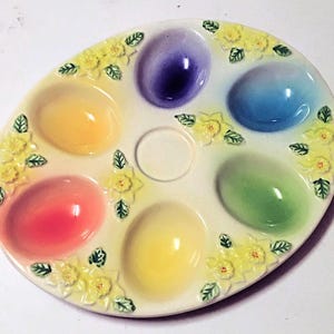 Könnte beinhalten: Ovale Keramik-Osterei-Servierschale mit sechs eiförmigen Vertiefungen in Gelb, Rot, Lila, Blau, Grün und Gelb. Die Schale ist mit gelben Blumen und grünen Blättern an den Rändern verziert.