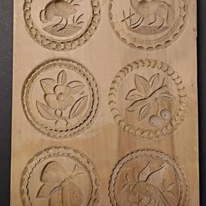 Könnte beinhalten: Eine Vintage-Keksform aus Holz mit sechs kreisförmigen, geschnitzten Designs. Die Designs umfassen einen Hahn, einen Fuchs, eine Blume, Blätter mit Beeren, einen Schmetterling und einen Vogel mit Blättern. Die Form ist aus hellem Holz gefertigt.