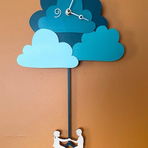 May include: A teal blue and light blue cloud-shaped clock with a pendulum featuring a silhouette of two people riding a bicycle. The clock face has a simple design with silver hands and a white swirl.