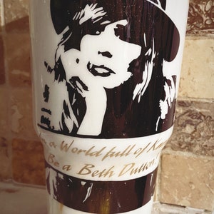 May include: White tumbler with a black and white silhouette of a woman wearing a hat. The text "In a world full of Kayce's, Be a Beth Dutton." is printed in gold on the tumbler.
