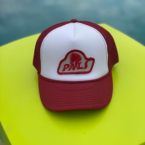 Può includere: Un cappello da camionista bianco e marrone con un logo ricamato rosso e bianco che dice "PALS" con un'immagine di un copricapo nativo americano all'interno della parola.
