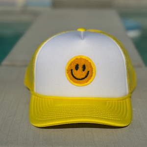 Könnte beinhalten: Eine gelbe und weiße Trucker-Kappe mit einem gelben Smiley-Gesicht, das auf der Vorderseite gestickt ist.