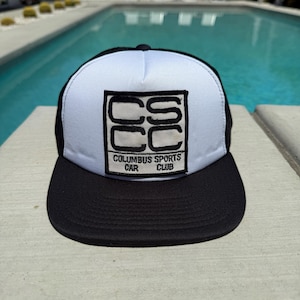 May include: A black and white trucker hat with a flat brim. The front panel is white with a black square logo that reads "CSCC" and "Columbus Sports Car Club". The hat is sitting on a concrete surface.