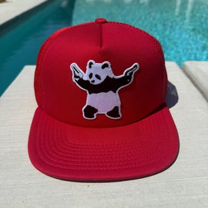May include: A red trucker hat with a white and black panda bear patch. The hat has a curved brim and a mesh back. The panda bear is depicted with its arms outstretched. The hat is sitting on a concrete surface.