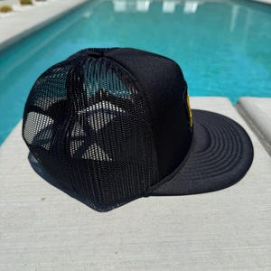 May include: Black trucker hat with a mesh back and a curved brim. The hat features a yellow patch on the front and a structured crown. The hat is sitting on a concrete surface next to a swimming pool.
