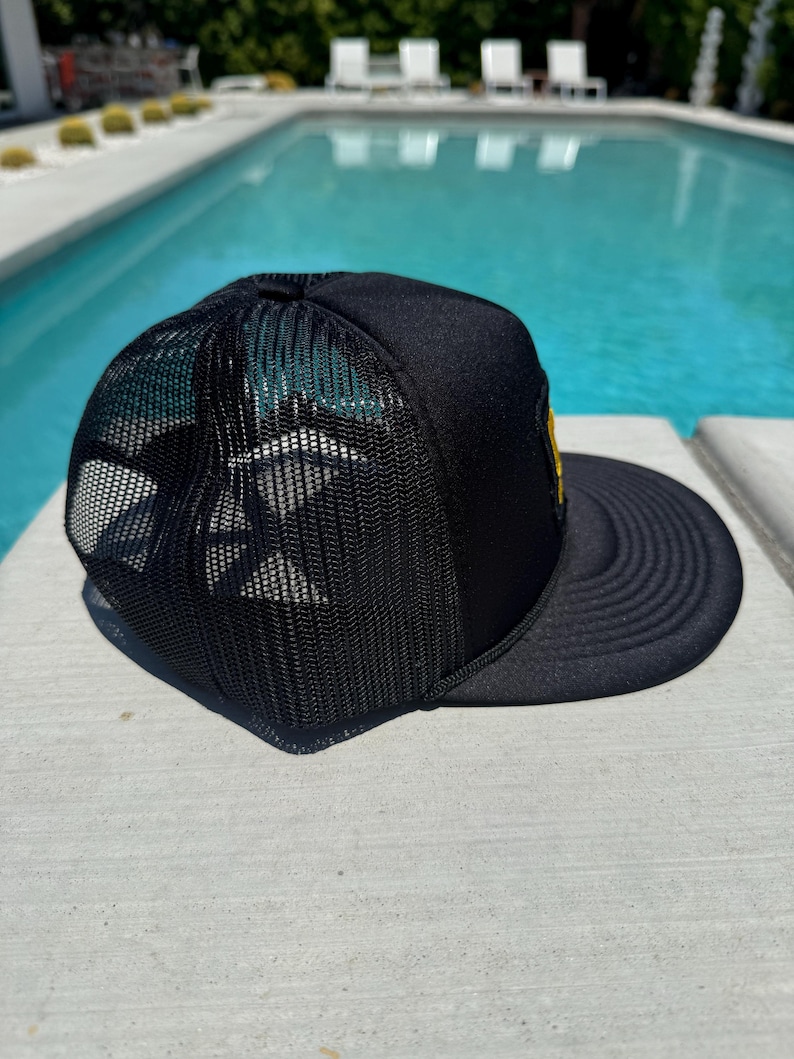 May include: Black trucker hat with a mesh back and a curved brim. The hat features a yellow patch on the front and a structured crown. The hat is sitting on a concrete surface next to a swimming pool.