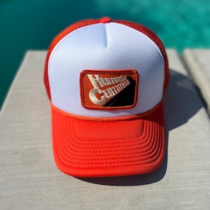 Peut inclure: Un chapeau de camionneur blanc et orange avec un patch noir et orange qui dit "Hot Rod Cummins".