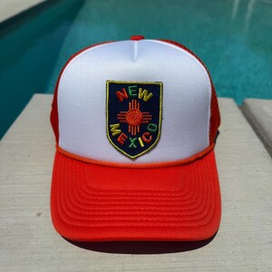 May include: A red and white trucker hat with a mesh back. The front panel features a patch with the words "NEW MEXICO" and a Zia symbol. The hat has a red brim and a white front panel.