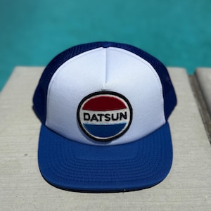 May include: A blue, white, and navy blue trucker hat with the word "DATSUN" in a circular patch. The hat has a blue brim and a white front panel with a mesh back.