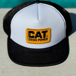 Pode incluir: Um chapéu de caminhoneiro branco e preto com um remendo amarelo que diz "CAT Diesel Power".