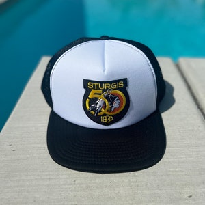 May include: A white and black trucker hat with a black mesh back. The front of the hat features a yellow and orange patch with the text "STURGIS 50 1990" and a Native American design.